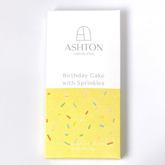 A Birthday Cake with Sprinkles chocolate bar box. Half of the box is white the other half is yellow with rainbow sprinkles.